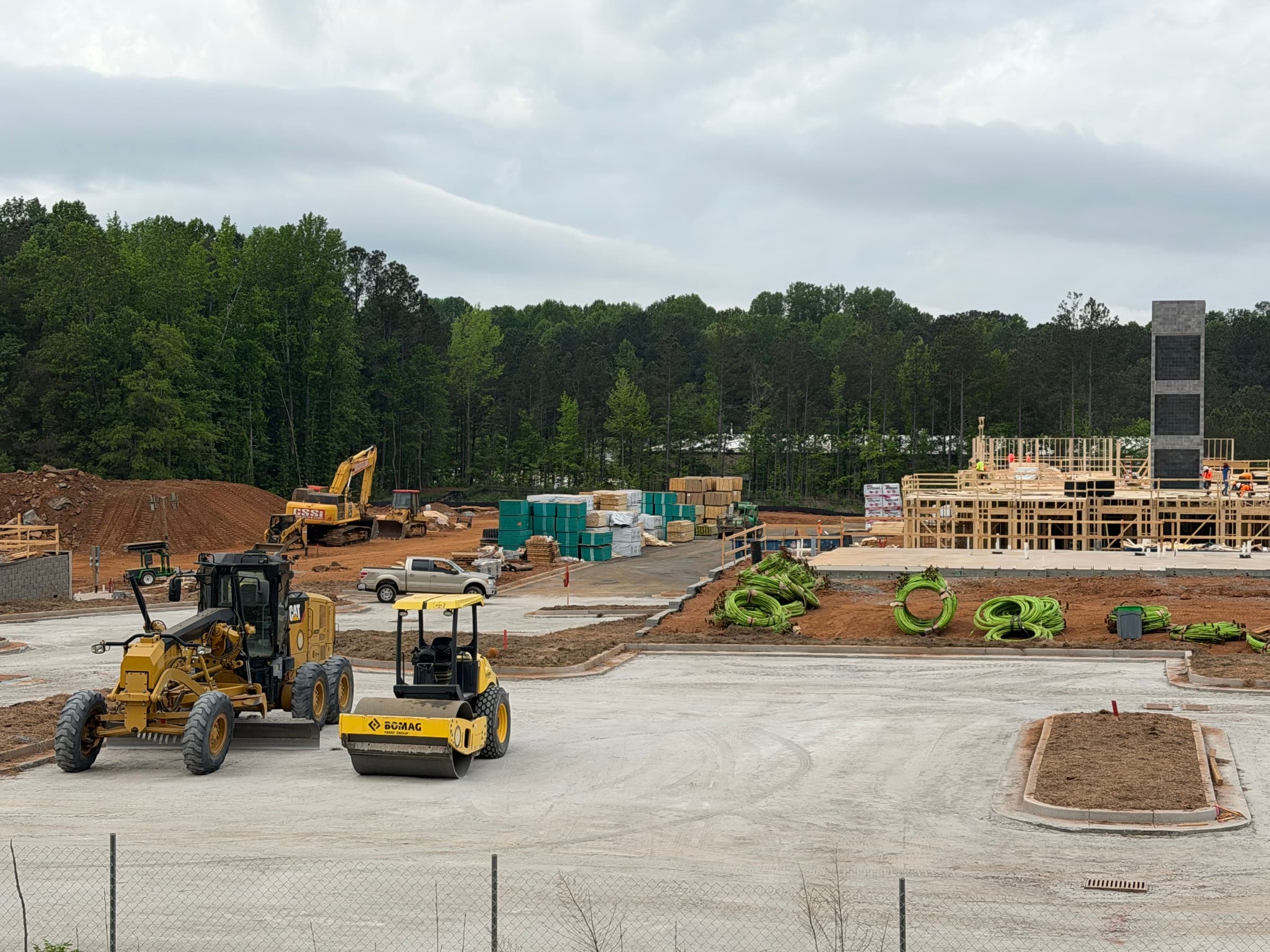 Motor grader and roller shaping a commercial pad while framing and utility materials are staged behind them.