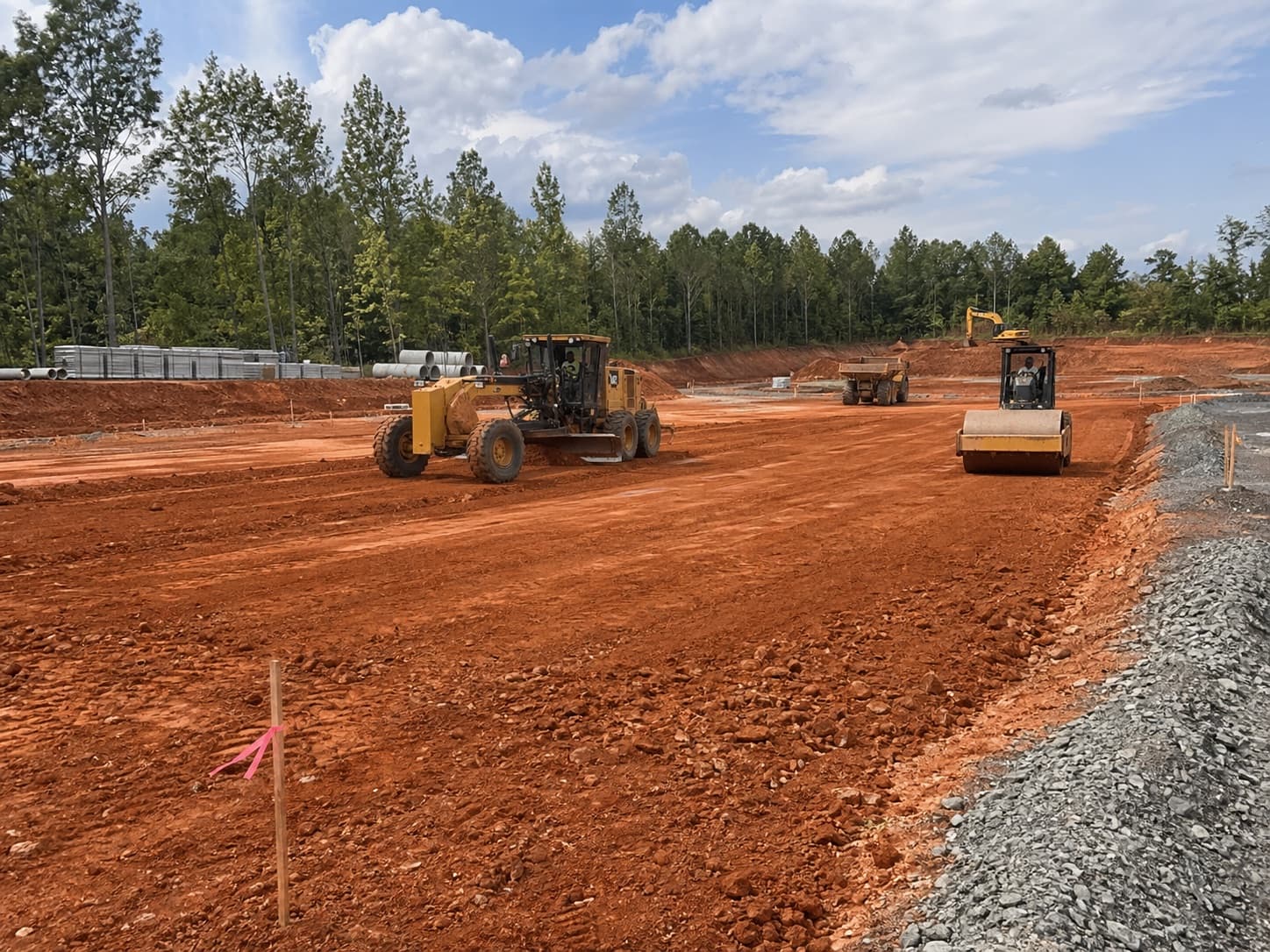 Motor grader, roller, and excavator grading a large red clay commercial pad.