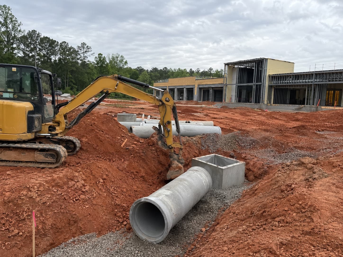 Excavator installing storm drainage pipe beside a framed commercial building on Georgia red clay.