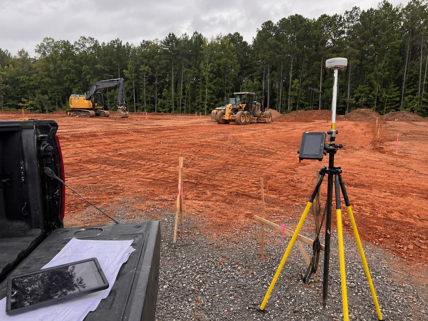 Survey tripod, plans, and machine-control layout equipment set up on a red clay commercial pad.