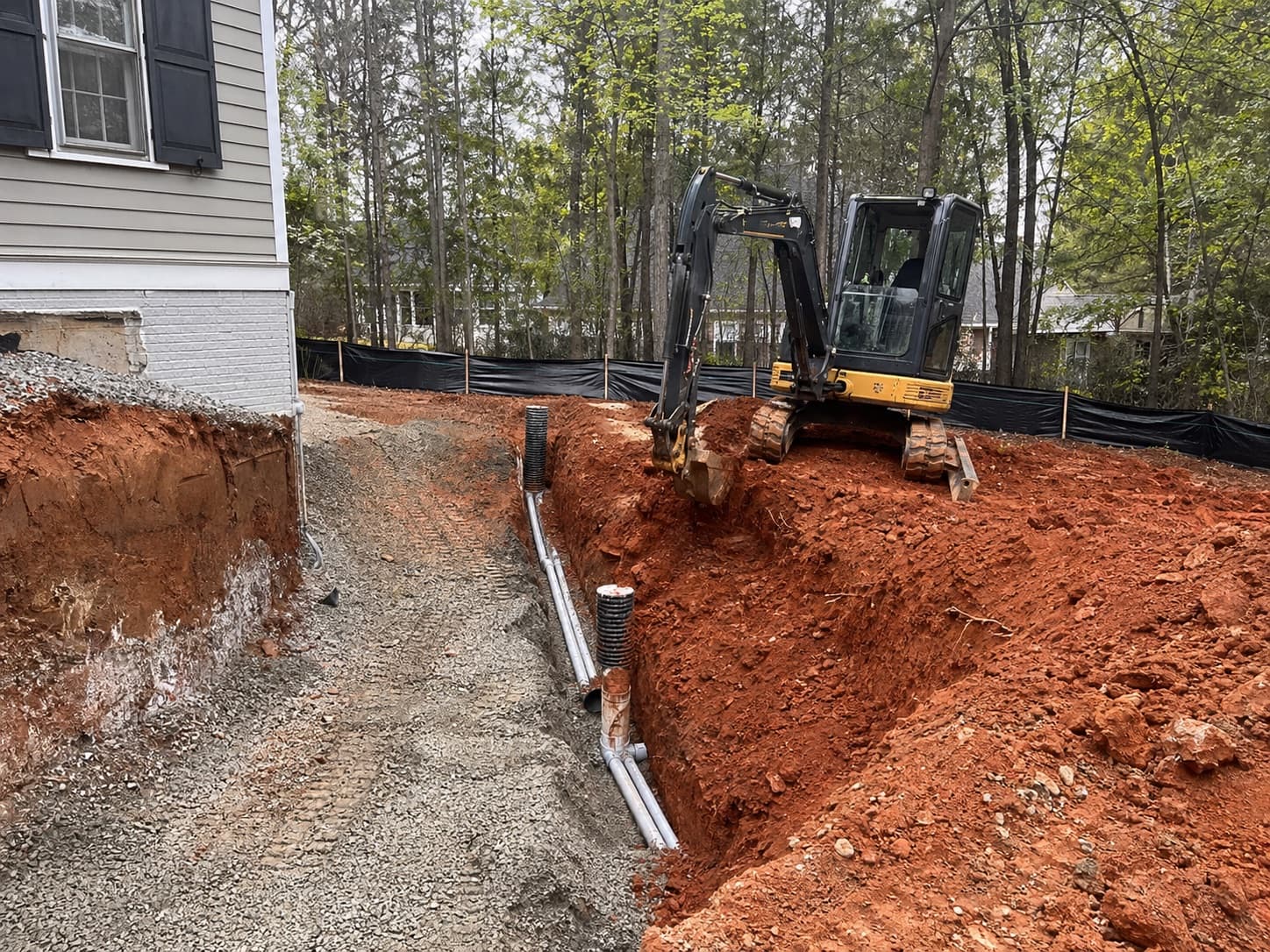Compact excavator trenching beside a home addition with red clay excavation and utility sleeves.