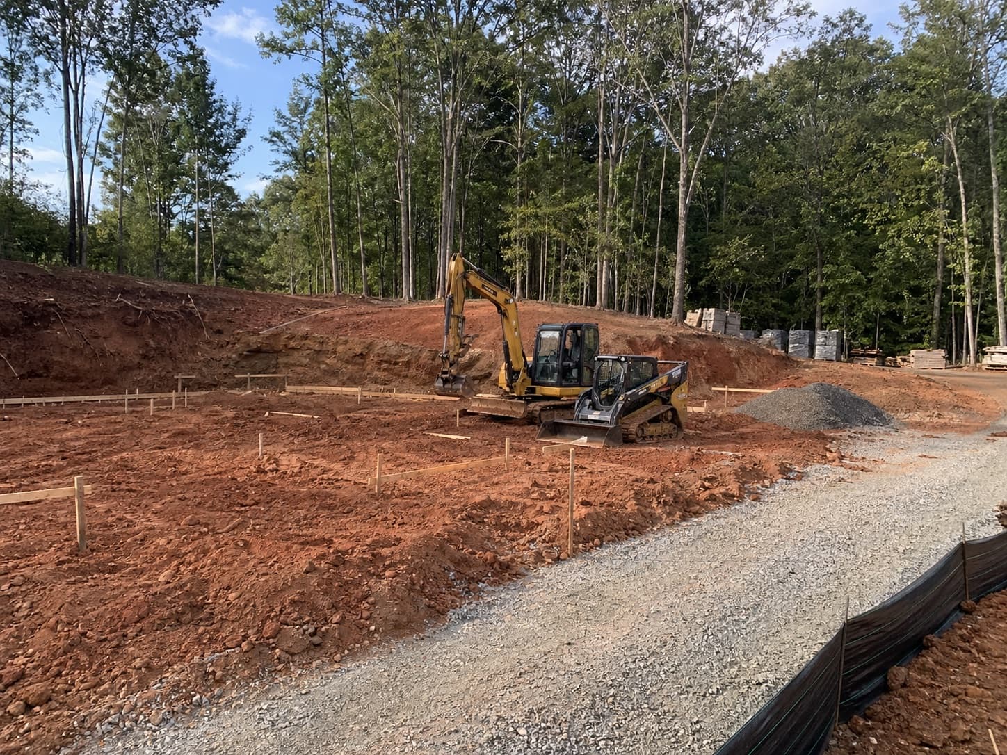 Compact excavator and skid steer preparing foundation staking on a wooded Georgia homesite.