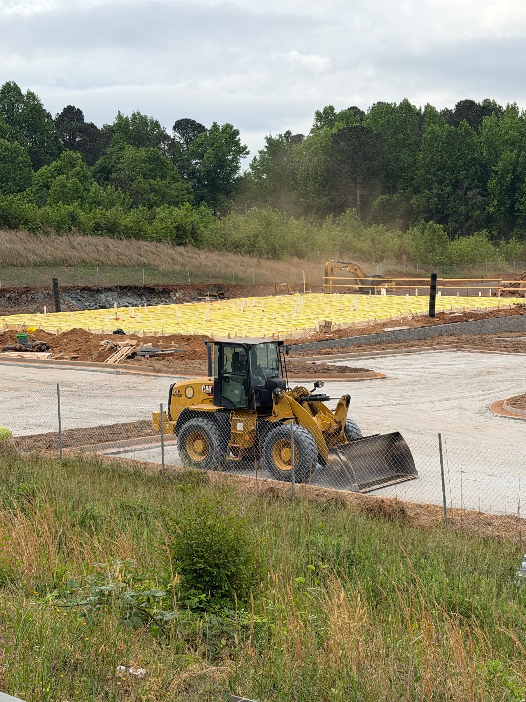 Retaining wall, slab prep, and commercial sitework along a wooded edge with active equipment in the background.